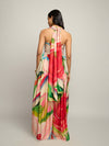 A woman viewed from the back wearing a halter-neck maxi dress with a vibrant abstract floral print in pink, green, blue, and cream, set against a plain light gray studio background.